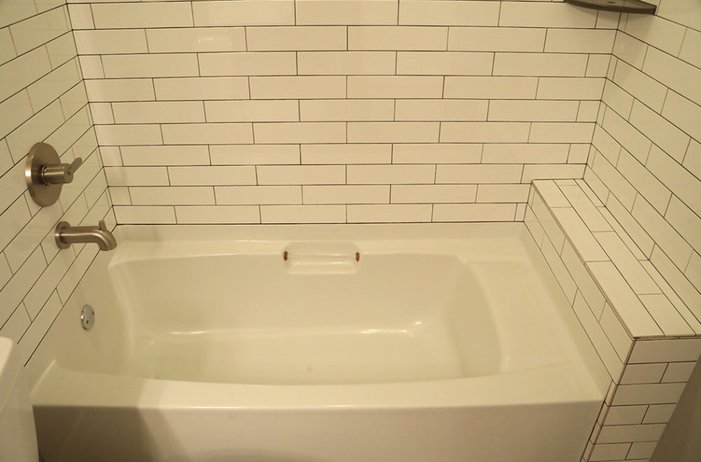 A close-up of a built-in bathtub with a beige tile surround in a subway tile pattern. The tub has a slight curve and features a wall-mounted faucet.