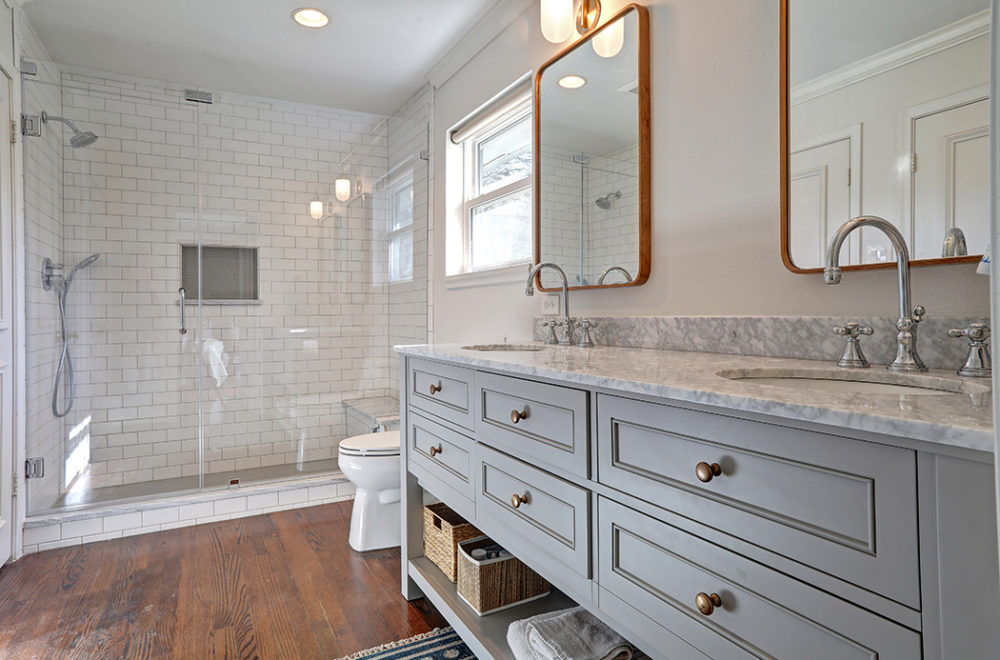 A bathroom with a gray vanity, brass handles, a large mirror with two rectangular faucets, and a wooden floor.