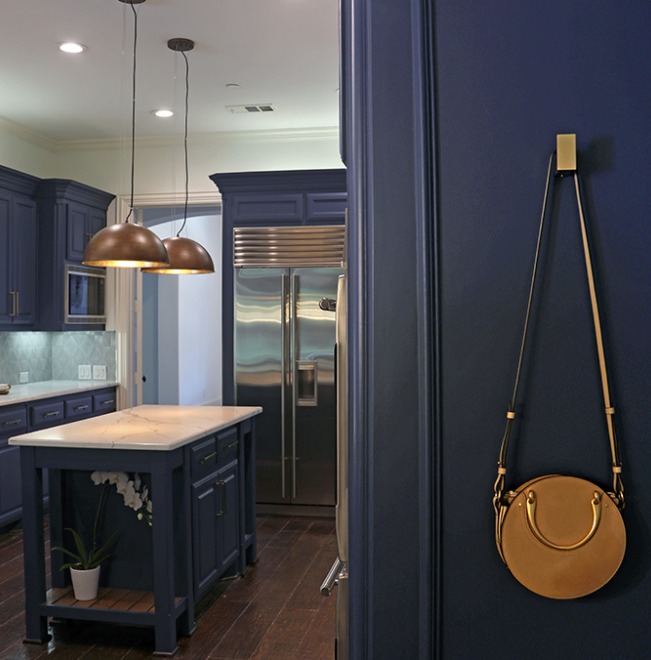 A side view of a kitchen showing a blue cabinet with a brass handle, a dark countertop, and a large copper pendant light. In the background, there's a glimpse of a stainless steel refrigerator and a built-in microwave.